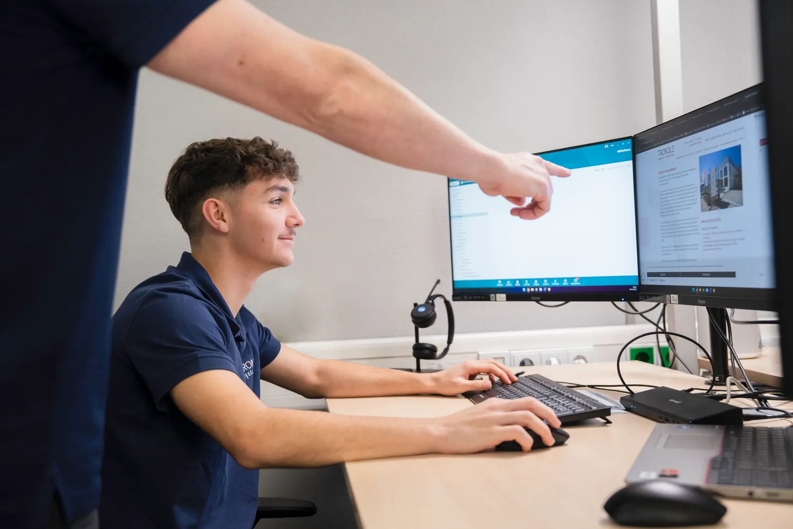 Ein junger Kollege am Arbeitsplatz, ein Kollege zeigt ihm etwas auf dem Bildschirm